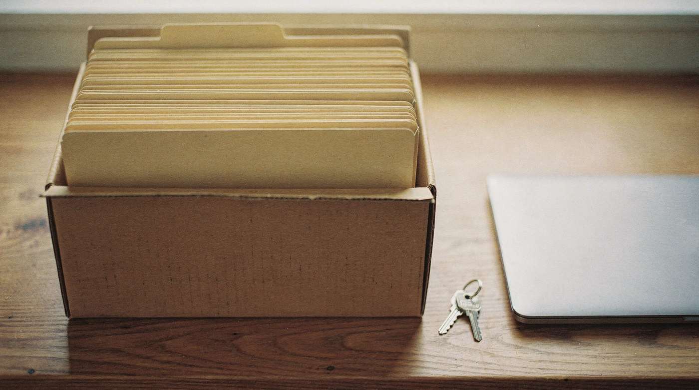 A plain records box filled with folders on a desk beside a laptop and keys